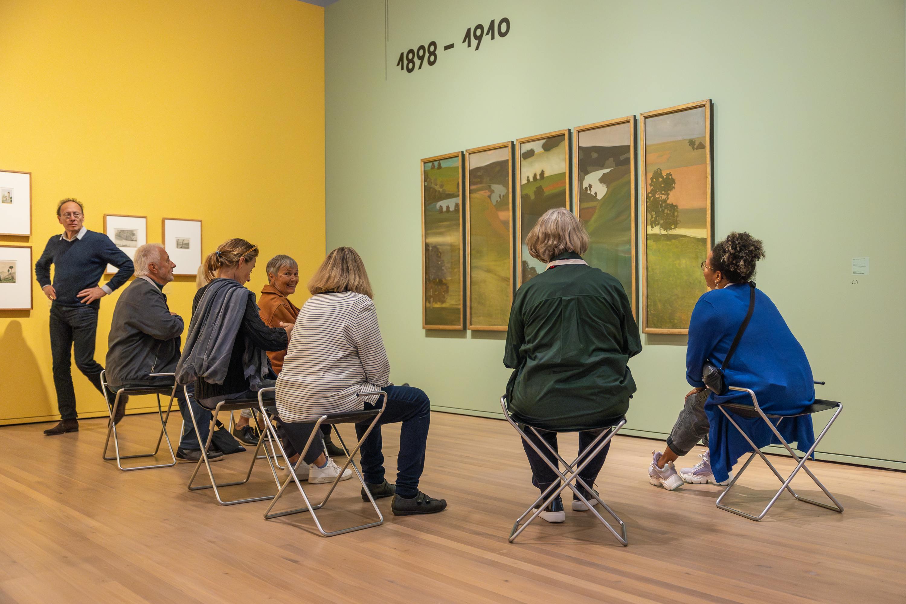 Eine Gruppe von 7 Personen sitzt nahe um ein Werk von Paul Klee und spricht darüber. und